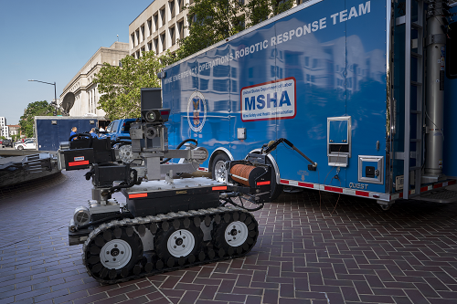 A robot with tracks like a bulldozer and a variety of mechanical parts sits outside a large blue trailer with MSHA&apos;s logo and &quot;Mine Emergency Operations Robotic Response Team&quot; 