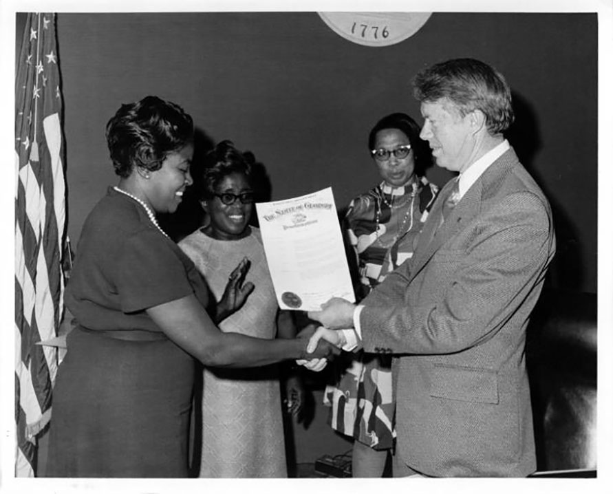 President Carter shakes Dorothy Bolden&apos;s hand and hands her a large piece of paper. Two other Black women look on in the background.