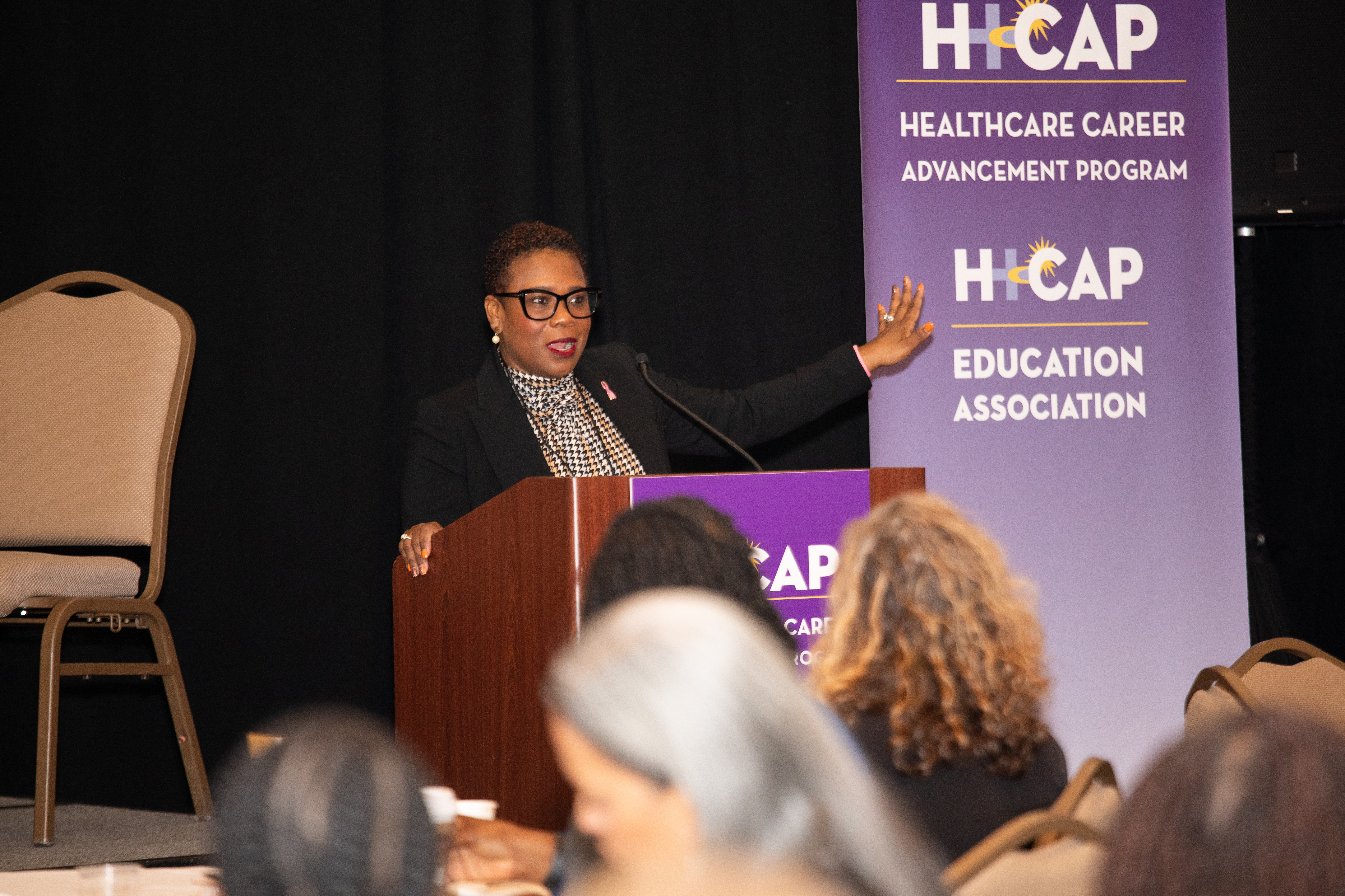 A woman in a dark jacket and glasses speaks at a podium. A banner in the background reads &quot;H-CAP Healthcare Career Advancement Program. H-CAP Education Association.&quot;
