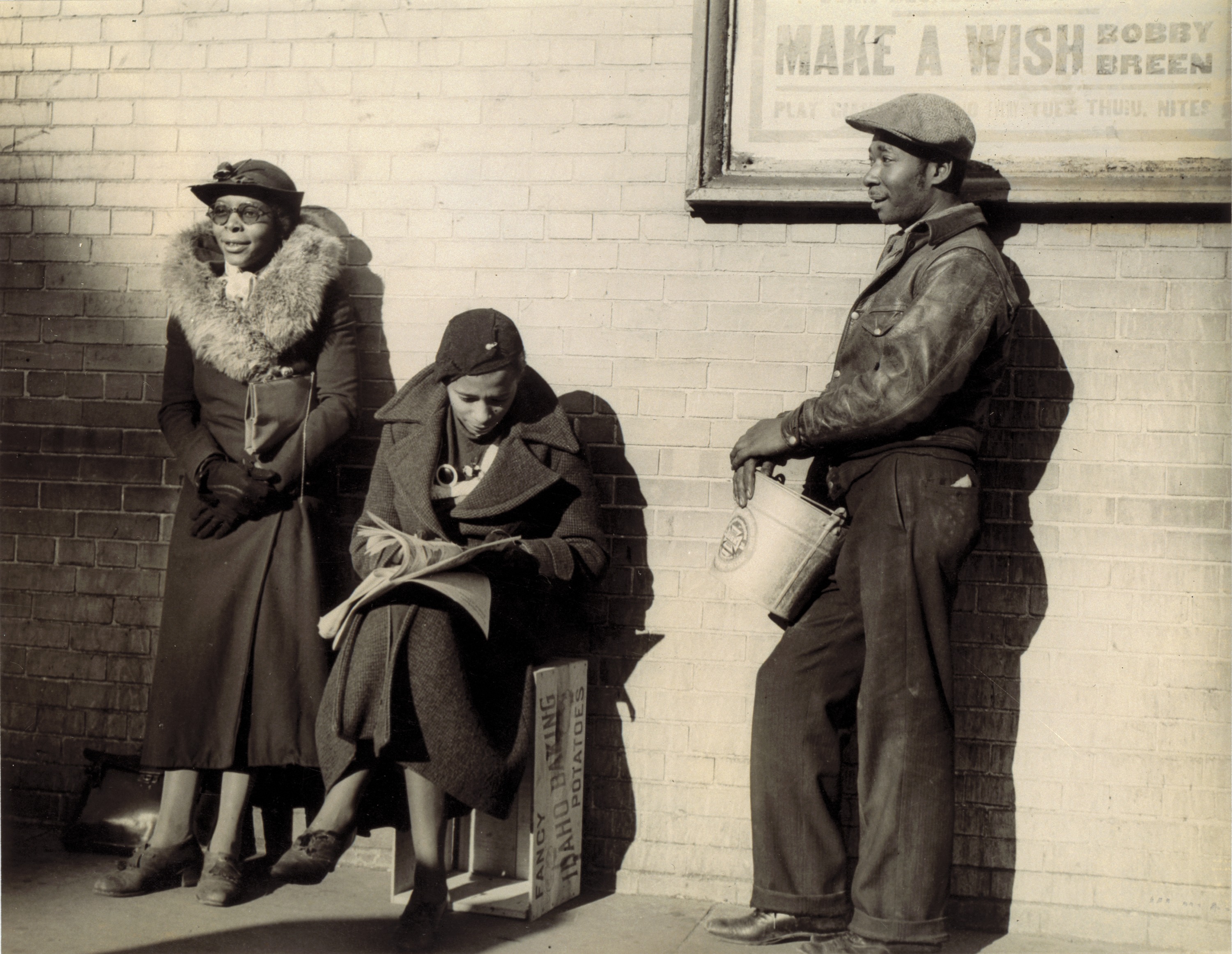 Black and white photograph showing two Black women and one man wearing coats and hats lean against a wall, waiting for employment. A movie poster behind them reads &quot;Make a Wish.&quot;