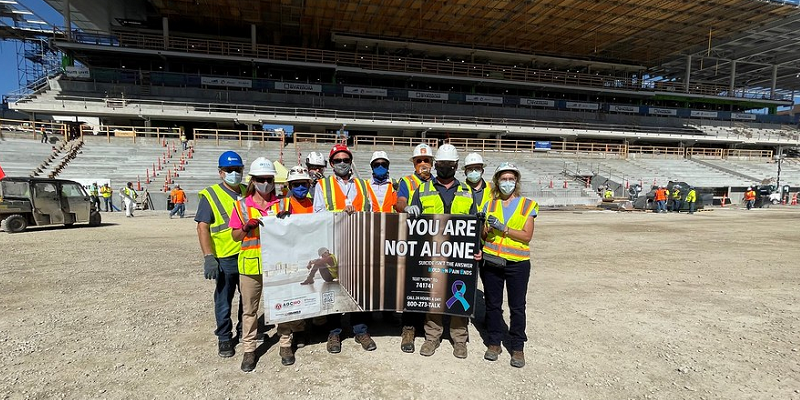 A group of construction workers standing in a stadium hold a banner that says &quot;You are not alone.&quot;