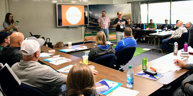 A group of safety trainers sit around tables in a classroom writing in notepads while they listen to a speaker. A screen behind the speaker has the image of a baseball with the words &quot;play ball.&quot;