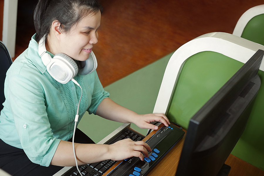 A young Asian person with a visual disability using a braille display to type. They are wearing a green blouse with white headphones around their neck.