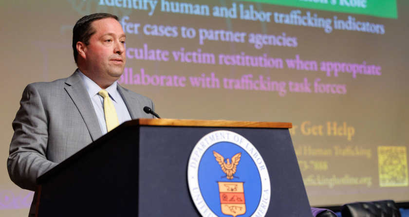 The image shows a person standing at a podium with the U.S. Department of Labor seal on it. The person is dressed in a suit and is delivering a speech. A presentation slide is visible in the background, related to human and labor trafficking topics.