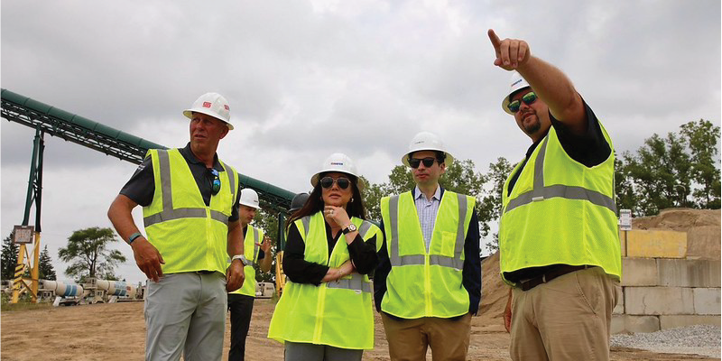 Keith Sonderling and Lori Chavez-DeRemer stand on a construction site with workers, all wearing safety vests and hard hats.