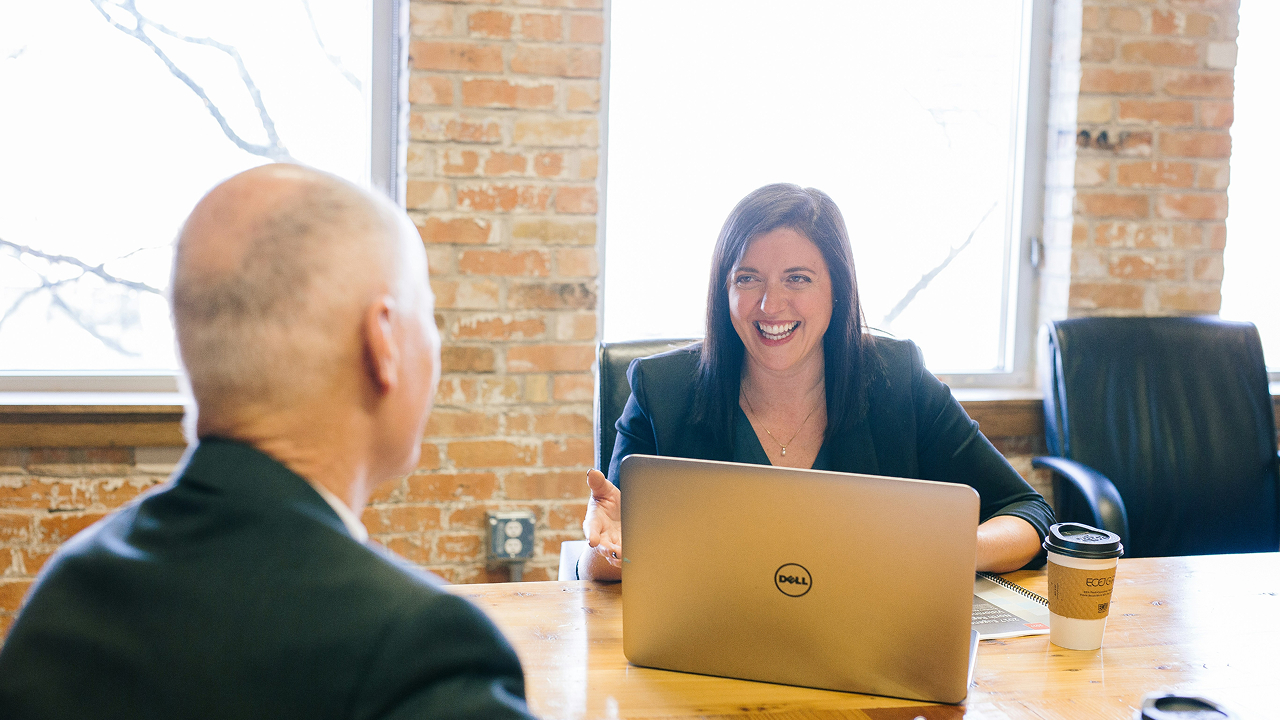 Two people sitting across a wooden table in an office, having a discussion with a laptop and a coffee cup on the table