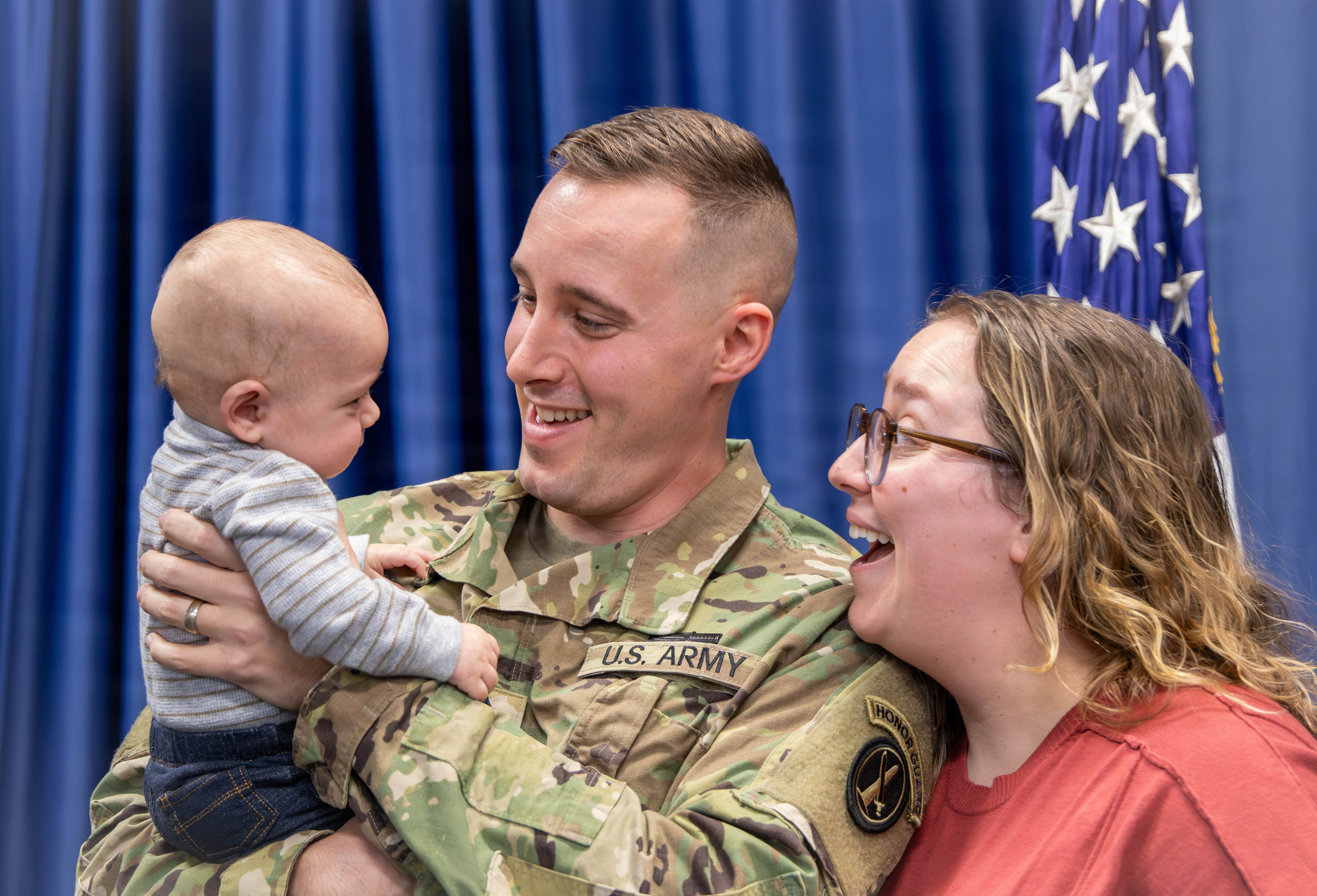 A U.S. Army soldier holds a baby while in uniform. He and his wife smile at the child.