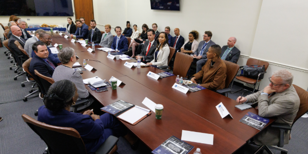 Dozens of people in professional clothing sit around a long table on which there are pamphlets and name placards.