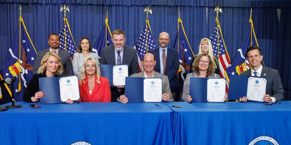 Ten people in professional clothes hold signed documents while posing for a photo. Flags of the United States and the Labor Department are visible behind them. 
