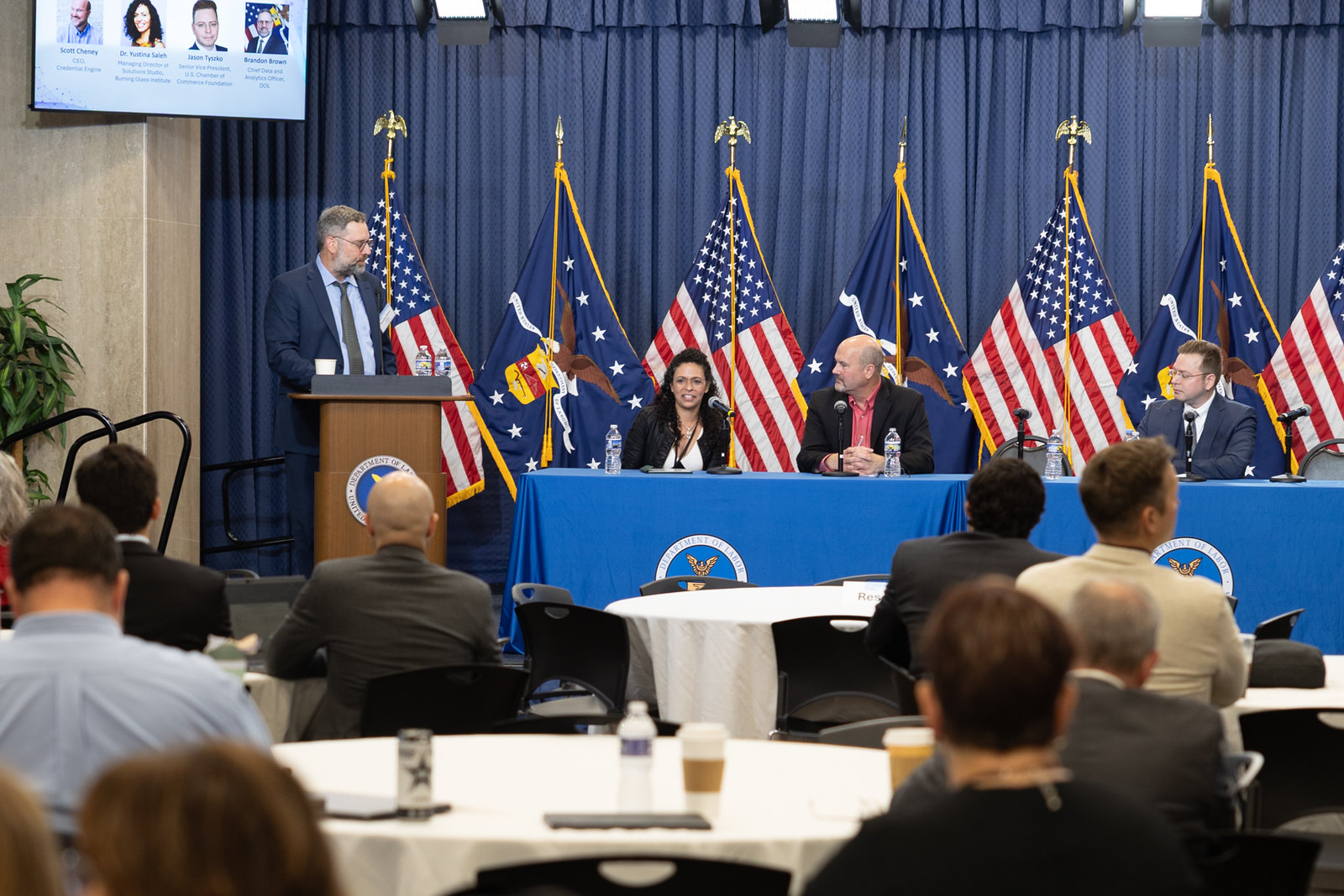 One person stands behind a podium and several sit at a table with microphones during a panel discussion at the U.S. Department of Labor. 