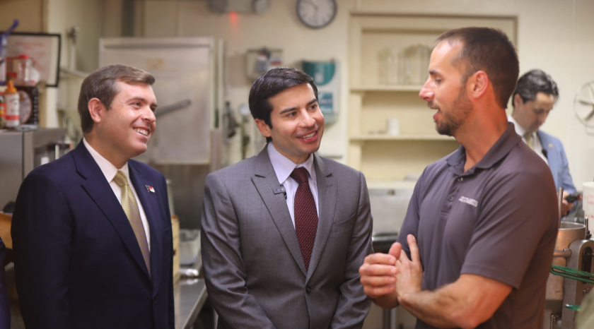 Acting Secretary of Labor stands in a workshop, chatting with two men - one in a suit, one in a collared shirt.