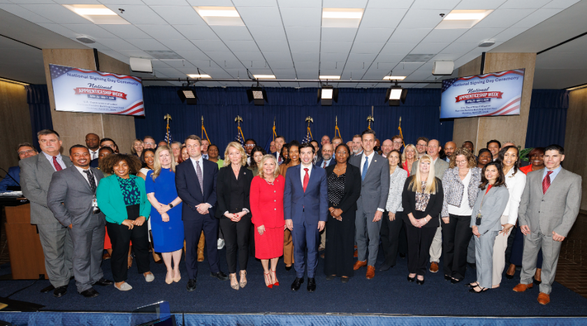 Dozens of men and women in professional clothing stand on a low stage, posing for a group photo. Signs behind them read “National Apprenticeship Week.” Acting Secretary of Labor Keith Sonderling is in the front row. 