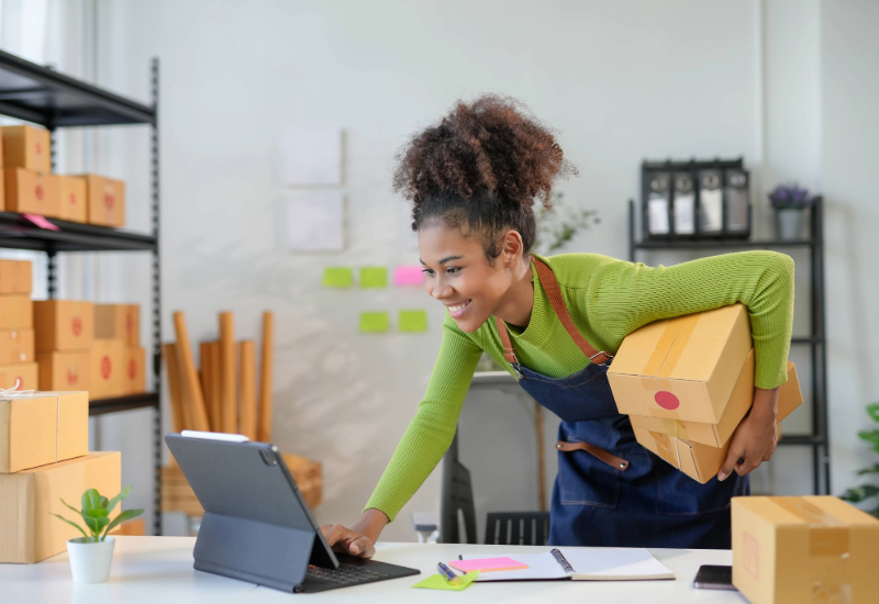 A young entrepreneur checks her laptop while holding shipping boxes.
