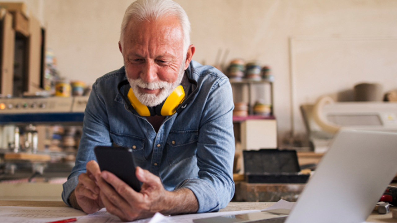 An old man in a workshop looking at a smartphone while working at a desk.