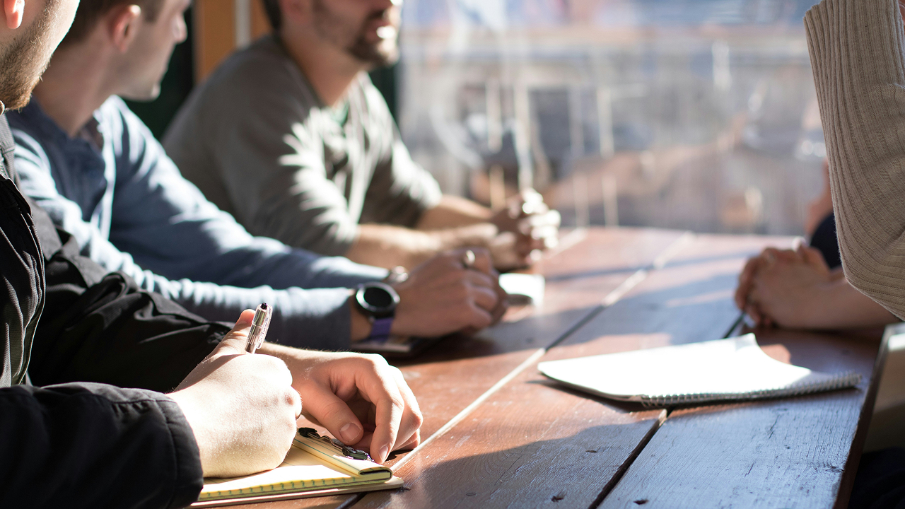 A group of people are working together at a table, and one person in the front is writing on a notepad.