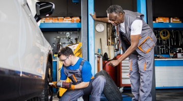 In a garage, a young man with Downs syndrome works on a car tire under the supervision of an adult mentor.