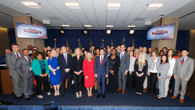 Dozens of men and women in professional clothing stand on a low stage, posing for a group photo. Signs behind them read “National Apprenticeship Week.” Acting Secretary of Labor Keith Sonderling is in the front row. 