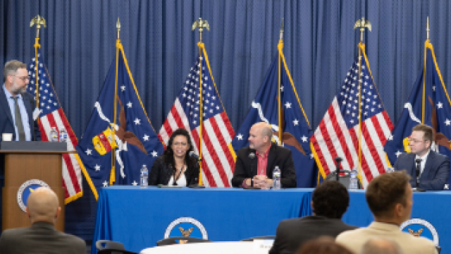 One person stands behind a podium and several sit at a table with microphones during a panel discussion at the U.S. Department of Labor. 