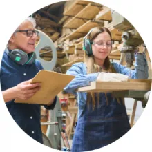An older worker supervises a younger one who is using a cutting tool
