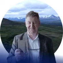 A man in business clothes holds a bar of nickel with Alaskan scenery in the background