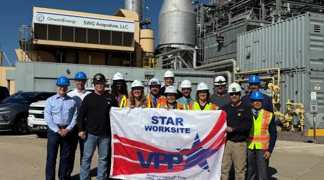 A group of people in hard hats stand in front of an energy plant on a sunny day. They hold a banner that says "VPP STAR Worksite."