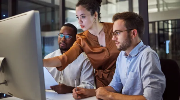 Three workers lean over a desk, looking at a computer screen.