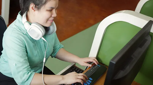 A young Asian person with a visual disability using a braille display to type. They are wearing a green blouse with white headphones around their neck.