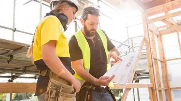 a student and teacher wearing yellow vests looking at blueprints at a construction site.
