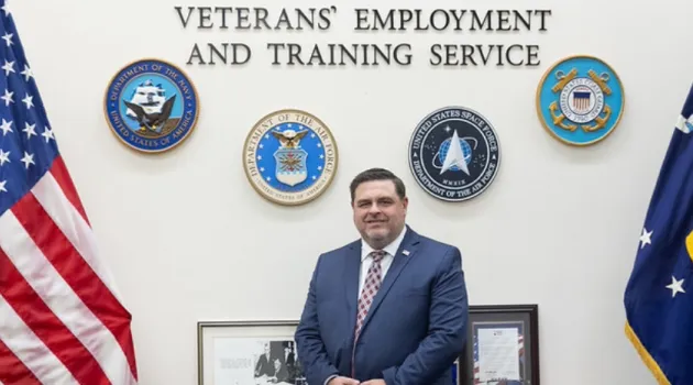 A man in a blue suit sits on a desk with framed pictures behind him. The military seals surround text reading Office of the Assistant Secretary Veterans’ Employment and Training Service on the wall. The US flag is on the left and the DOL flag is on the right.