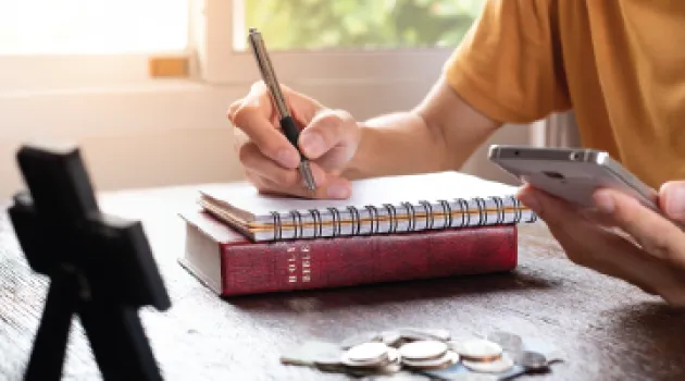 A person takes notes while using a calculator. A bible and cross are visible on the table.