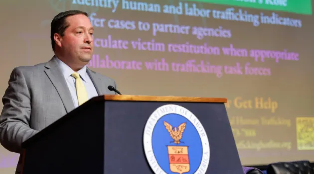 The image shows a person standing at a podium with the U.S. Department of Labor seal on it. The person is dressed in a suit and is delivering a speech. A presentation slide is visible in the background, related to human and labor trafficking topics.