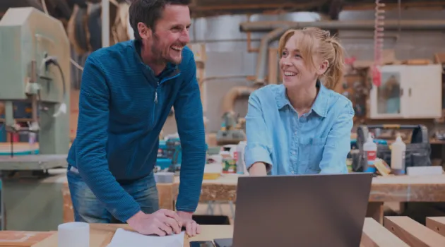 A man and a woman in a workshop are laughing and talking while looking at something on a laptop.