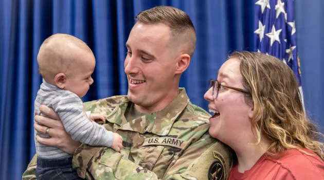 A U.S. Army soldier holds a baby while in uniform. He and his wife smile at the child.