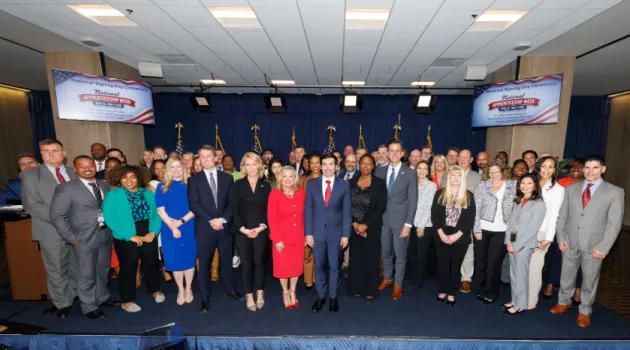 Dozens of men and women in professional clothing stand on a low stage, posing for a group photo. Signs behind them read “National Apprenticeship Week.” Acting Secretary of Labor Keith Sonderling is in the front row. 