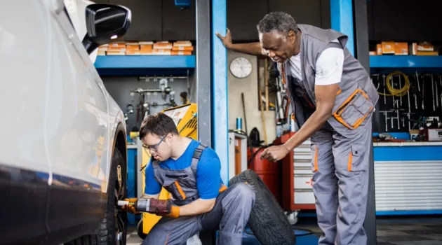 In a garage, a young man with Downs syndrome works on a car tire under the supervision of an adult mentor.