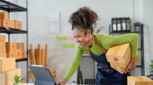 A young entrepreneur checks her laptop while holding shipping boxes.