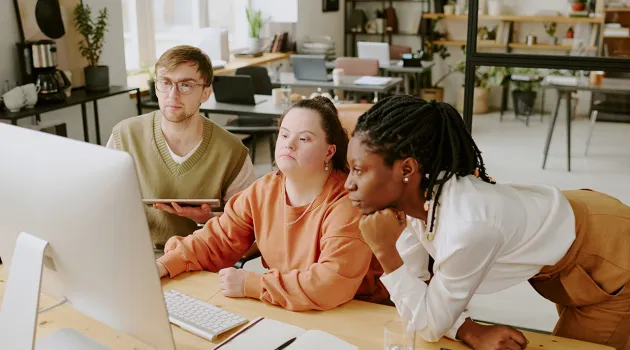Co-workers gather around a computer in a modern office. One has an apparent disability.