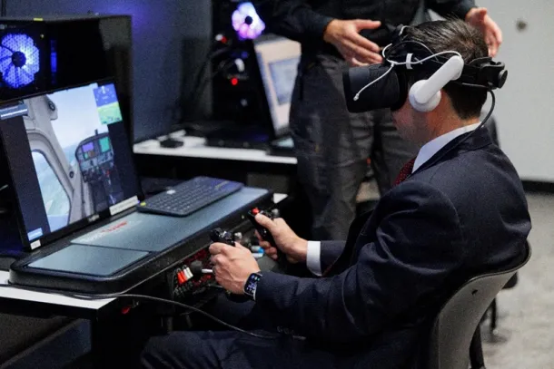 A man in a suit uses a virtual reality headset and aircraft controls as part of a pilot training simulation.