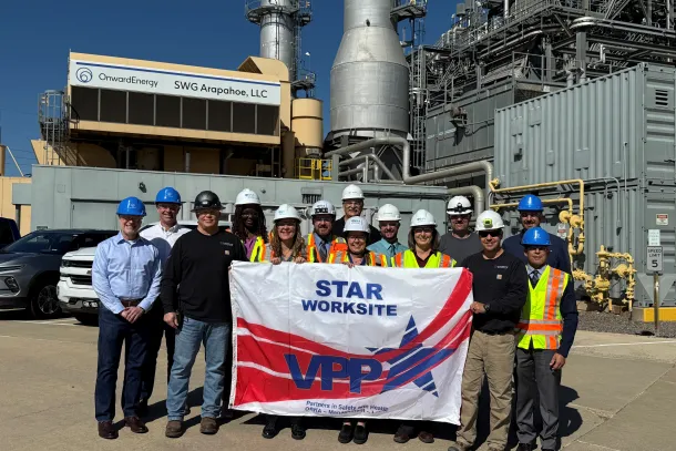 A group of people in hard hats stand in front of an energy plant on a sunny day. They hold a banner that says "VPP STAR Worksite."