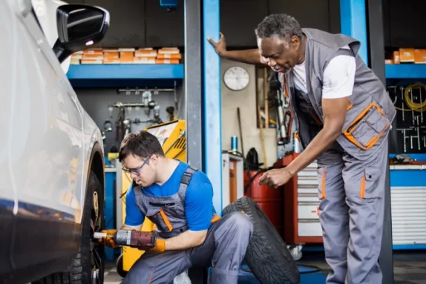 In a garage, a young man with Downs syndrome works on a car tire under the supervision of an adult mentor.