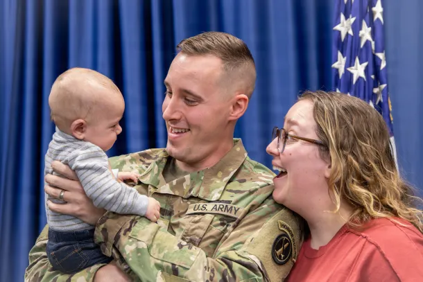 A U.S. Army soldier holds a baby while in uniform. He and his wife smile at the child.