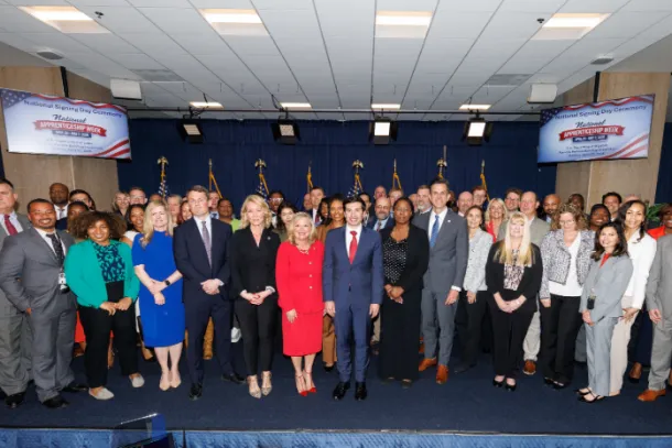 Dozens of men and women in professional clothing stand on a low stage, posing for a group photo. Signs behind them read “National Apprenticeship Week.” Acting Secretary of Labor Keith Sonderling is in the front row. 