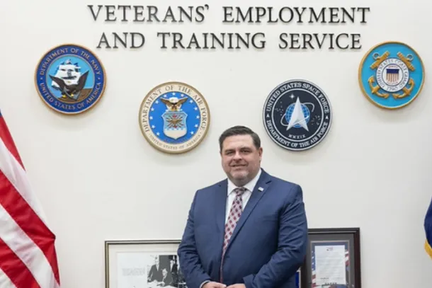 A man in a blue suit sits on a desk with framed pictures behind him. The military seals surround text reading Office of the Assistant Secretary Veterans’ Employment and Training Service on the wall. The US flag is on the left and the DOL flag is on the right.