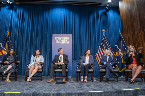 Six panelists sit in chairs on a stage. A Federal Tech Day banner and flags are behind them.