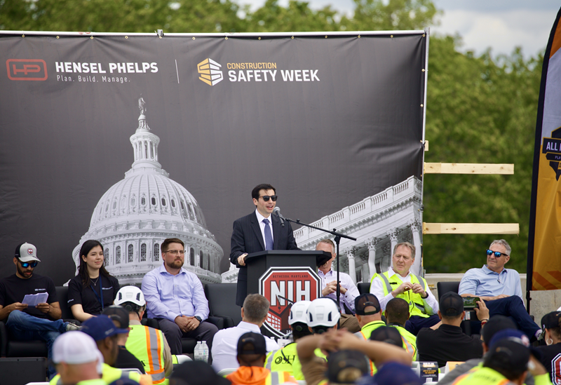 Deputy Secretary Keith Sonderling speaks at a construction safety event. A large backdrop behind him says "Hensel Phelps" and "Construction Safety Week." The podium has the National Institutes of Health logo.