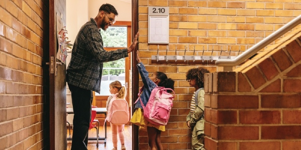 A teacher stands in the doorway of a classroom, giving high fives to young children as they enter.