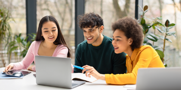 Three young people working with a laptop and notebooks at a table, smiling and engaged in their task. The setting appears to be a bright room with large windows.
