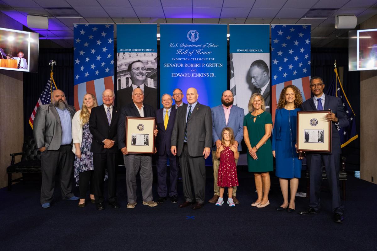 A group of family members and government officials attend a ceremony to induct Sen. Robert Griffin and Howard Jenkins Jr. into the Hall of Honor. Banners behind them have the American flag and black and white photos of both men.