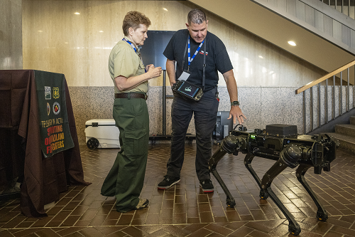 A man wears a virtual controller that directs a black Boston Dynamics robot dog while speaking with a Forest Service employee.