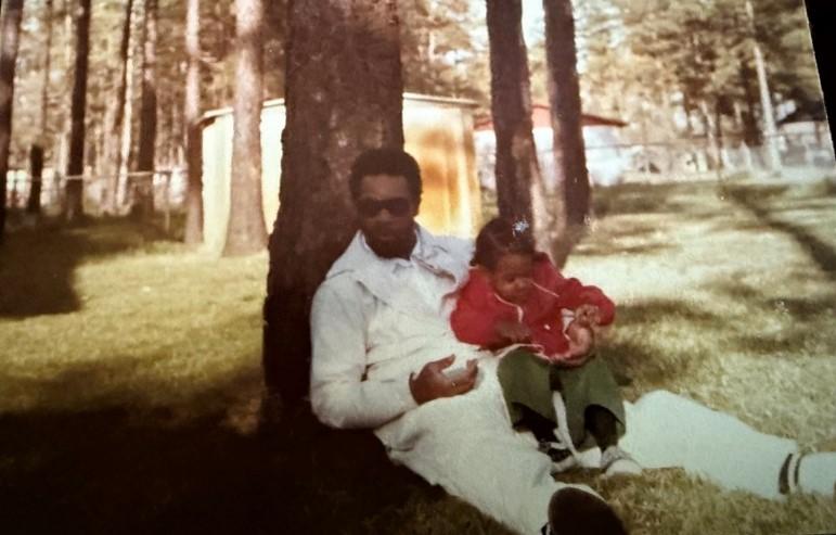 A man sits on the ground with his back against a tree, holding his young daughter.